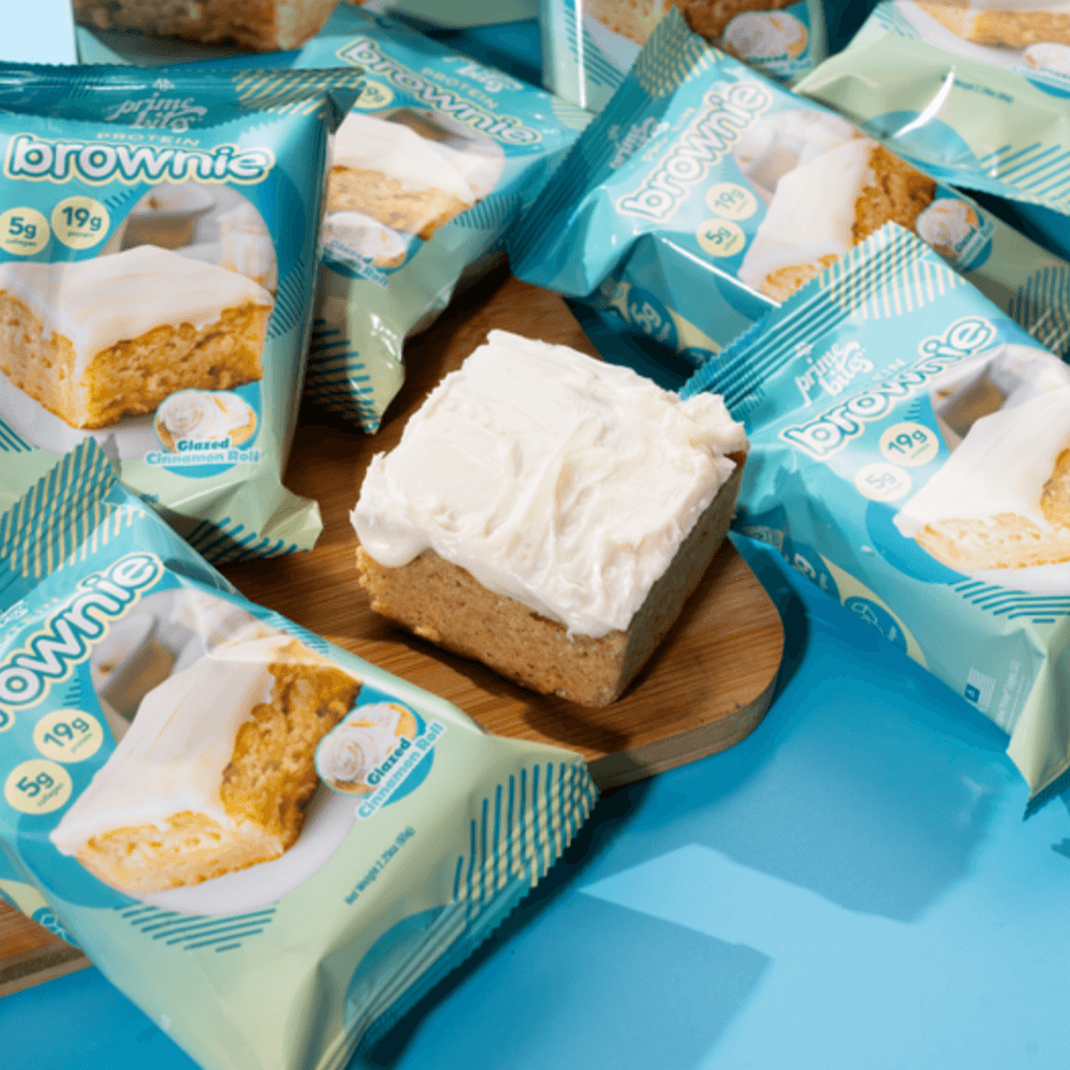 Glazed cinnamon roll brownie on a wooden board, surrounded by snack packaging, showcasing delicious protein treats.