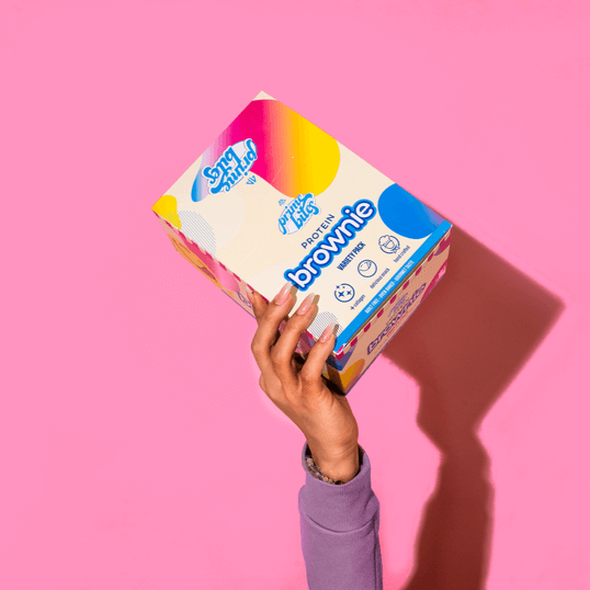 Hand holding Prime Bites brownie protein box with colorful packaging on a pink background.